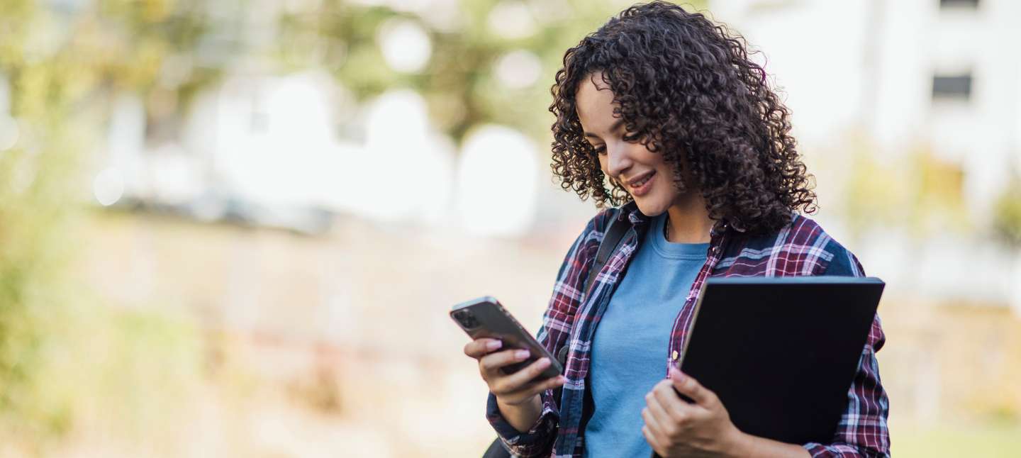 A college student walking on campus, carrying her laptop and using her phone while she walks and smiles at the phone.